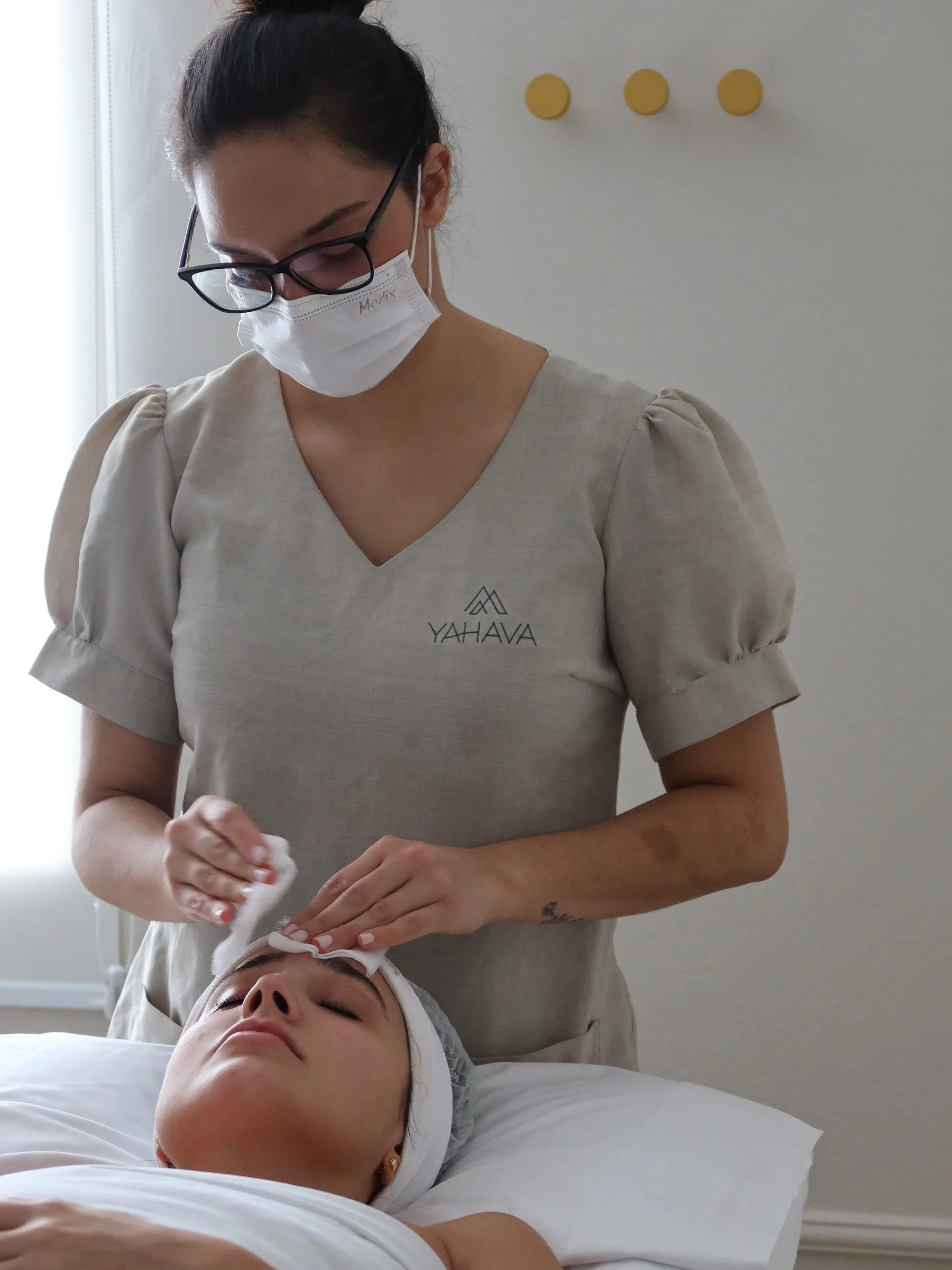 Esteticista da Yahava, de máscara e uniforme da casa, fazendo a limpeza de pele de uma cliente